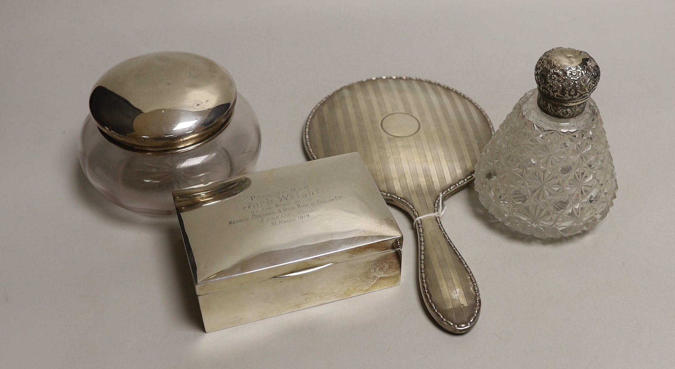 A George V silver backed hand mirror, a silver mounted cigarette box, a silver topped powder bowl and a silver topped scent bottle.